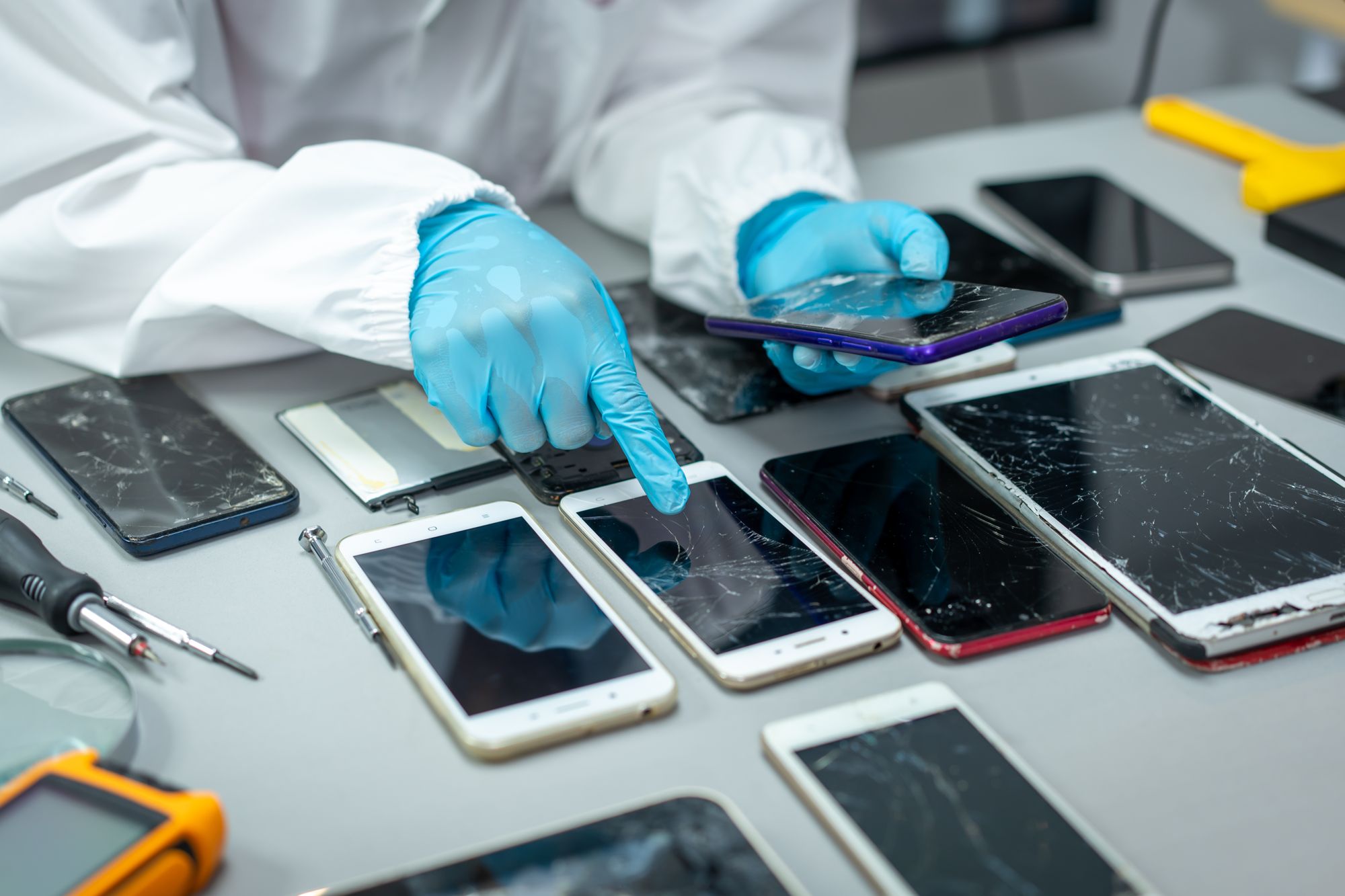 Gloved technician grading a table of returned smartphones with cracked screens